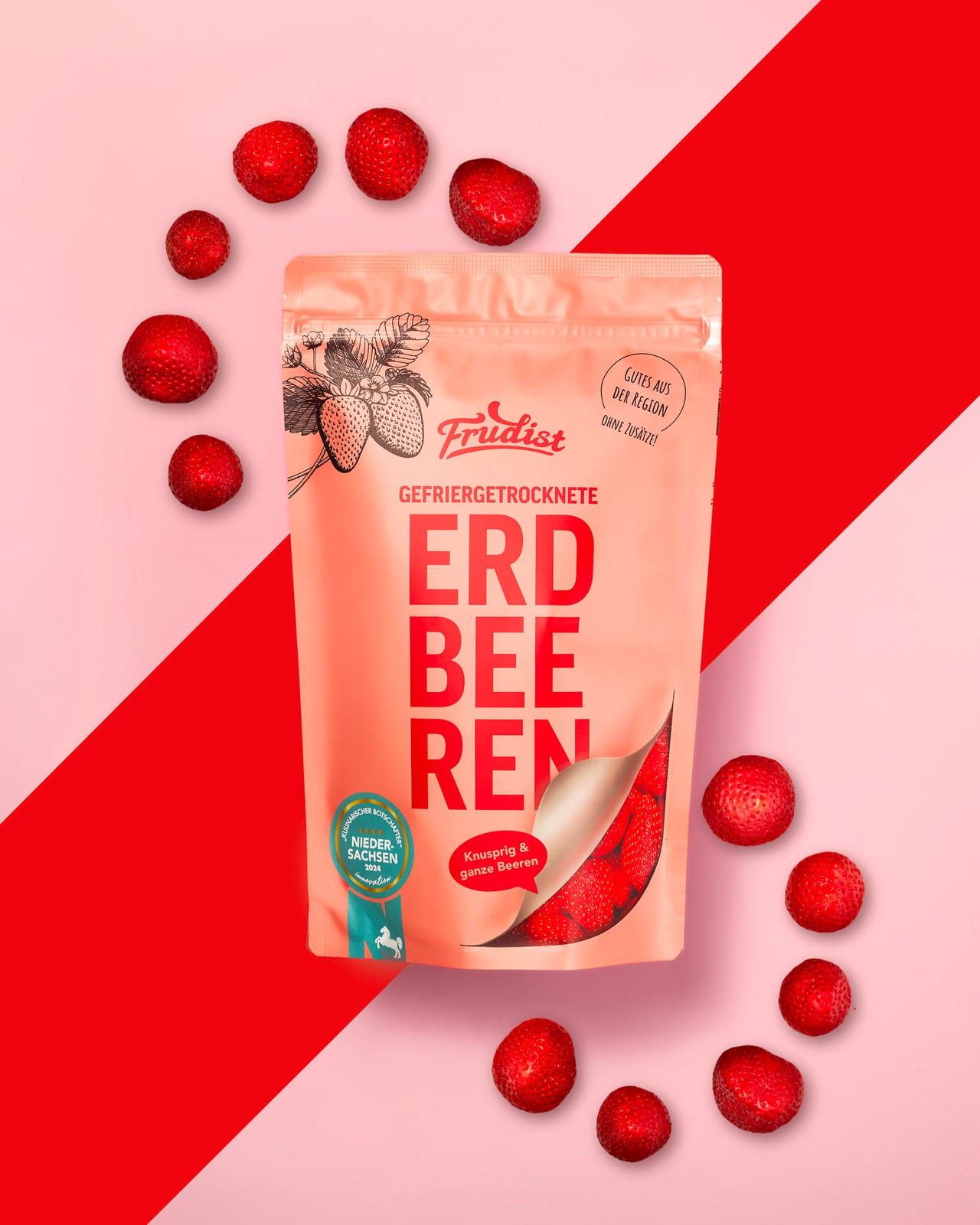 Frudist-Beutel mit gefriergetrockneten Erdbeeren auf Rosa und Rot, umrahmt von Erdbeeren