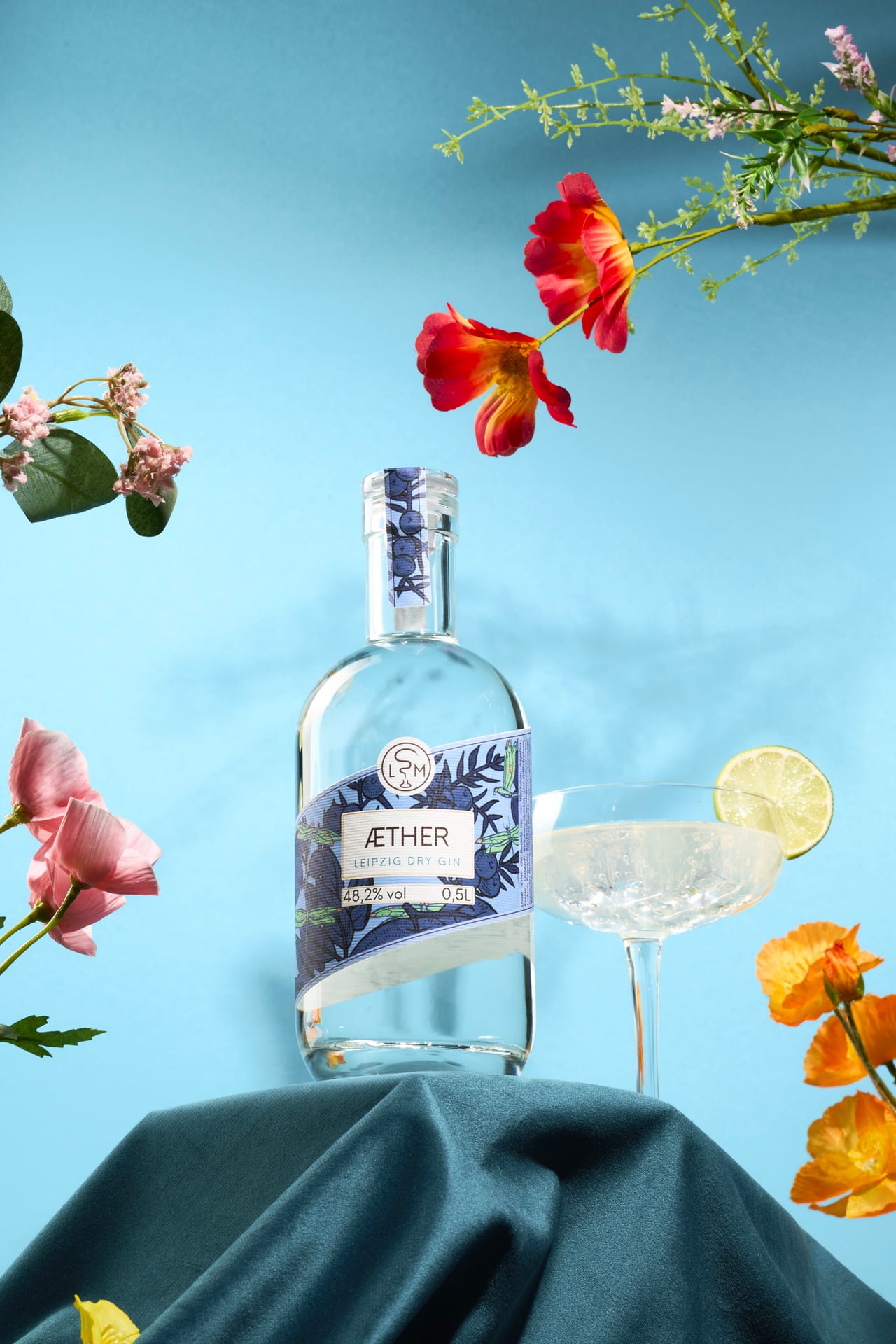 Äther-Gin-Flasche der Leipziger Spirituosenmanufaktur mit Cocktailglas und roten Blüten vor hellblauem Hintergrund