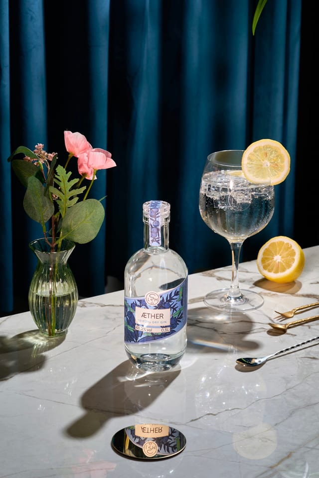 Äther-Gin-Flasche mit Tonic-Glas, Zitrone und rosa Blüten auf Marmor vor blauem Samtvorhang