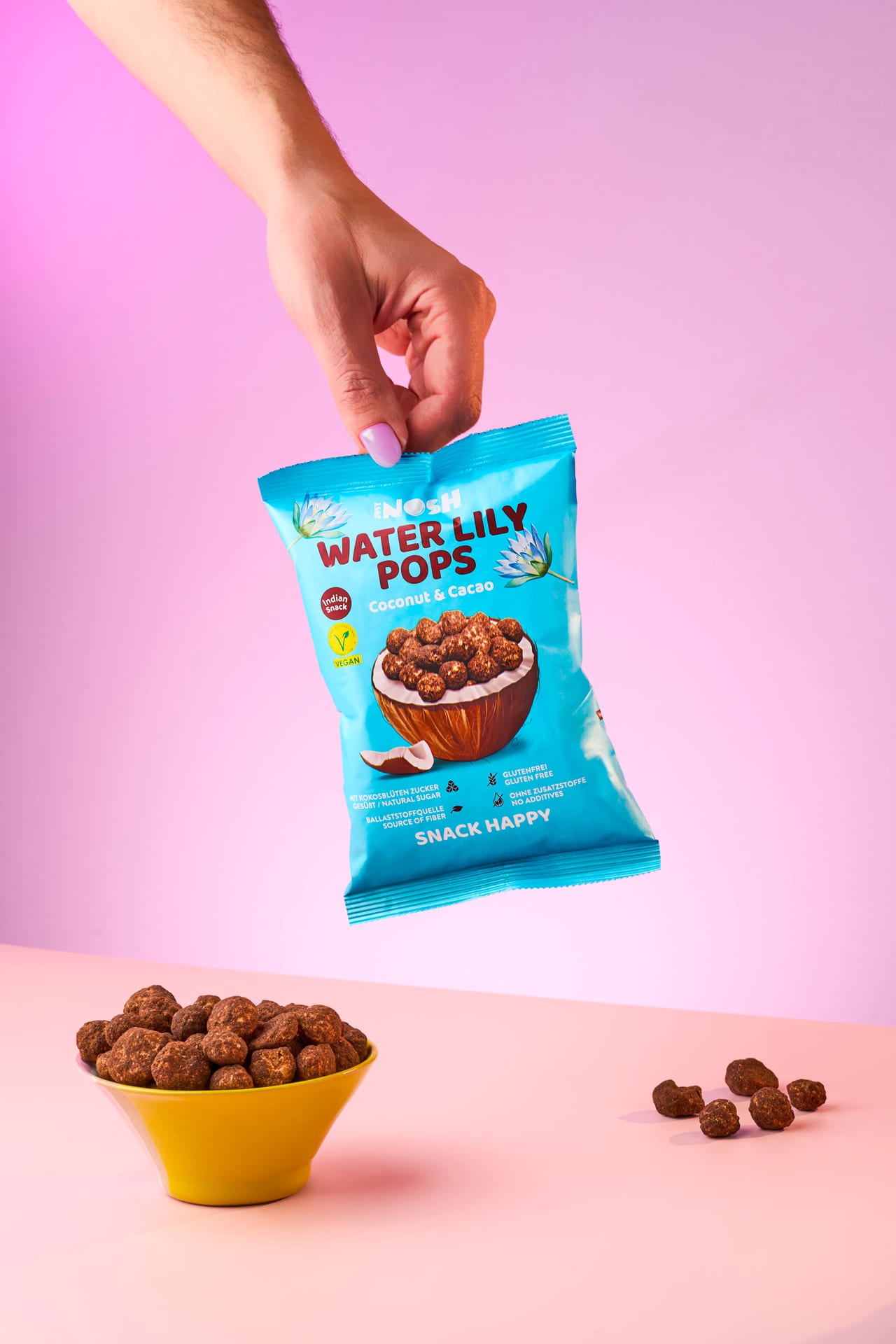 Hand hält Water Lily Pops Coconut & Cacao über einer gelben Schale vor rosa Hintergrund