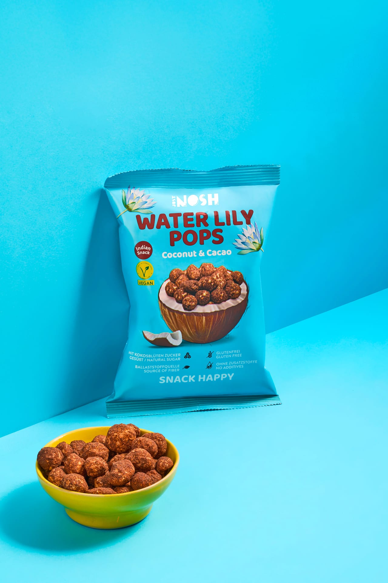 Water Lily Pops Coconut & Cacao Beutel und gelbe Schale mit Pops vor hellblauem Hintergrund